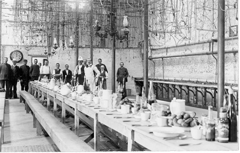 black and white images of a group of fourteen men standing at far end of a long table set for lunch