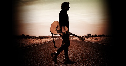 Man standing on a rad with an outback landscape in the background and he is holding a guitar