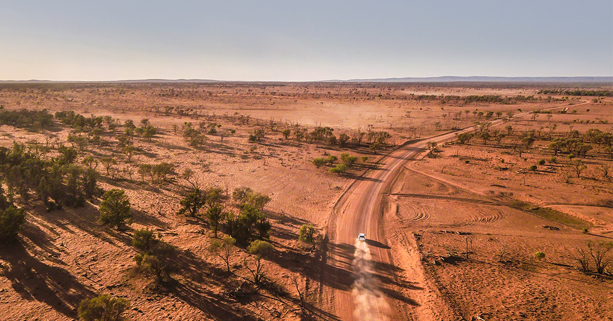 A car drives along a dirt road in an outback setting