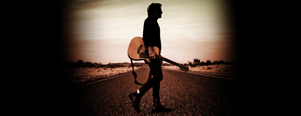 Ian-Moss-standing-on-a-highway-carrying-a-guitar.png