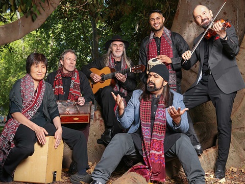 Group of six musicians, posing outdoors with their instruments. Each member is wearing a scarf with red and black patterns.