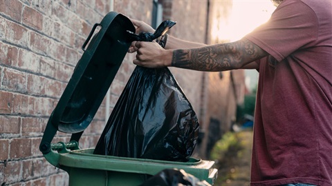 green garbage bin in front of brick home with a person placing a garbage bag inside.