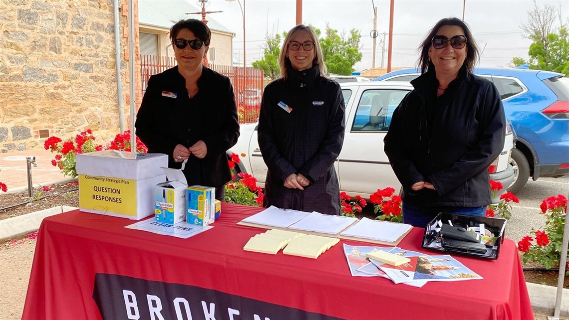 Three female council employees, standing behind a table at a local community consultation session with brochures